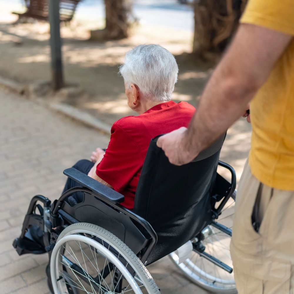 A caregiver pushing a senior woman in a wheelchair along a sunny path